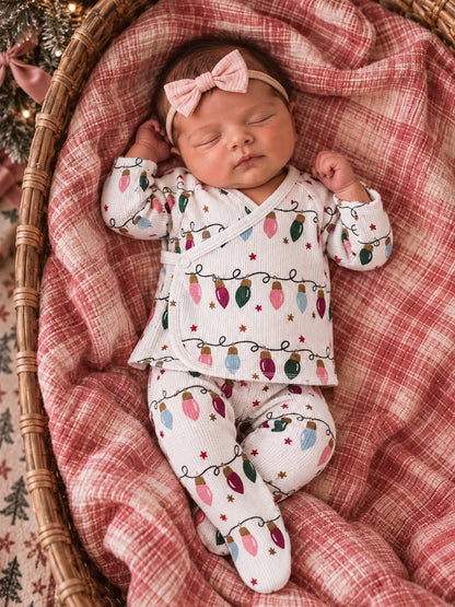 Sleeping baby in festive outfit with holiday lights; nestled in a cozy basket on a plaid blanket.