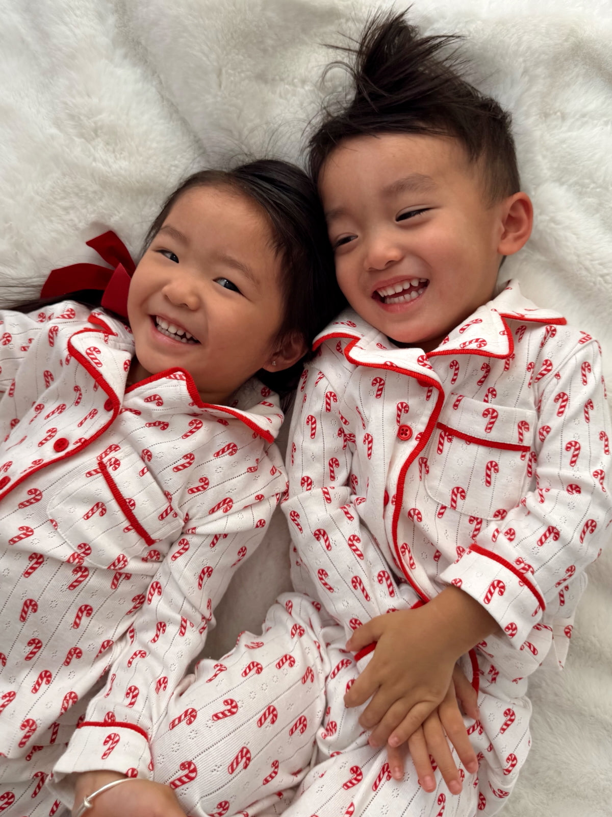 Two smiling children in festive pajamas with candy cane print, lying together on a soft blanket.