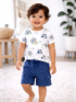 Smiling toddler in a soccer-themed shirt and shorts, standing on a soft carpeted floor.