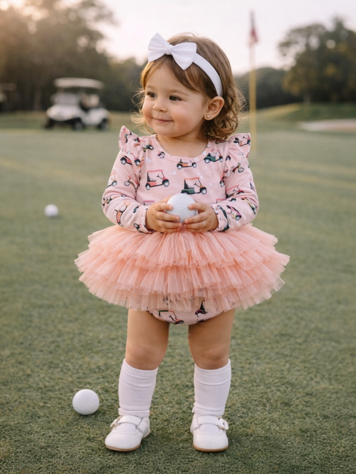 Young girl in a pink dress holding a golf ball on a green course, smiling with a white headband. Golf cart in background.