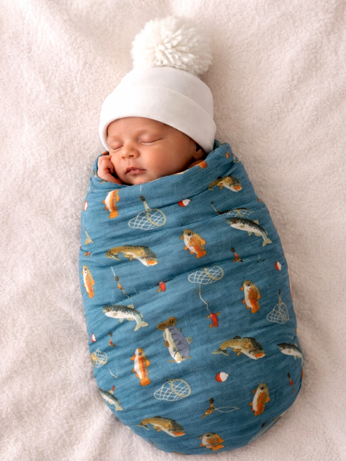 Newborn wrapped in a fish-patterned blanket wearing a white knit hat, sleeping peacefully on a soft surface.
