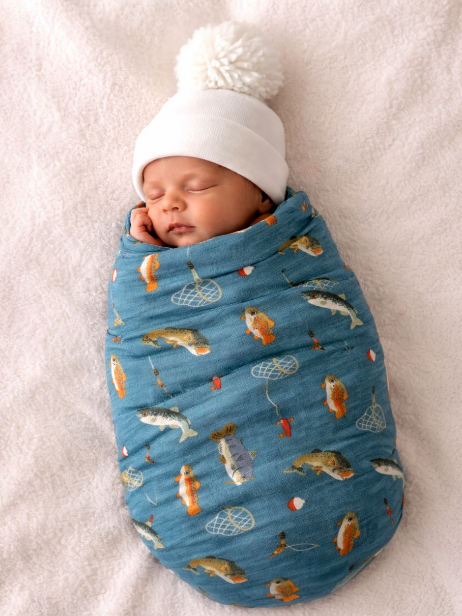 Newborn wrapped in a fish-patterned blanket wearing a white knit hat, sleeping peacefully on a soft surface.
