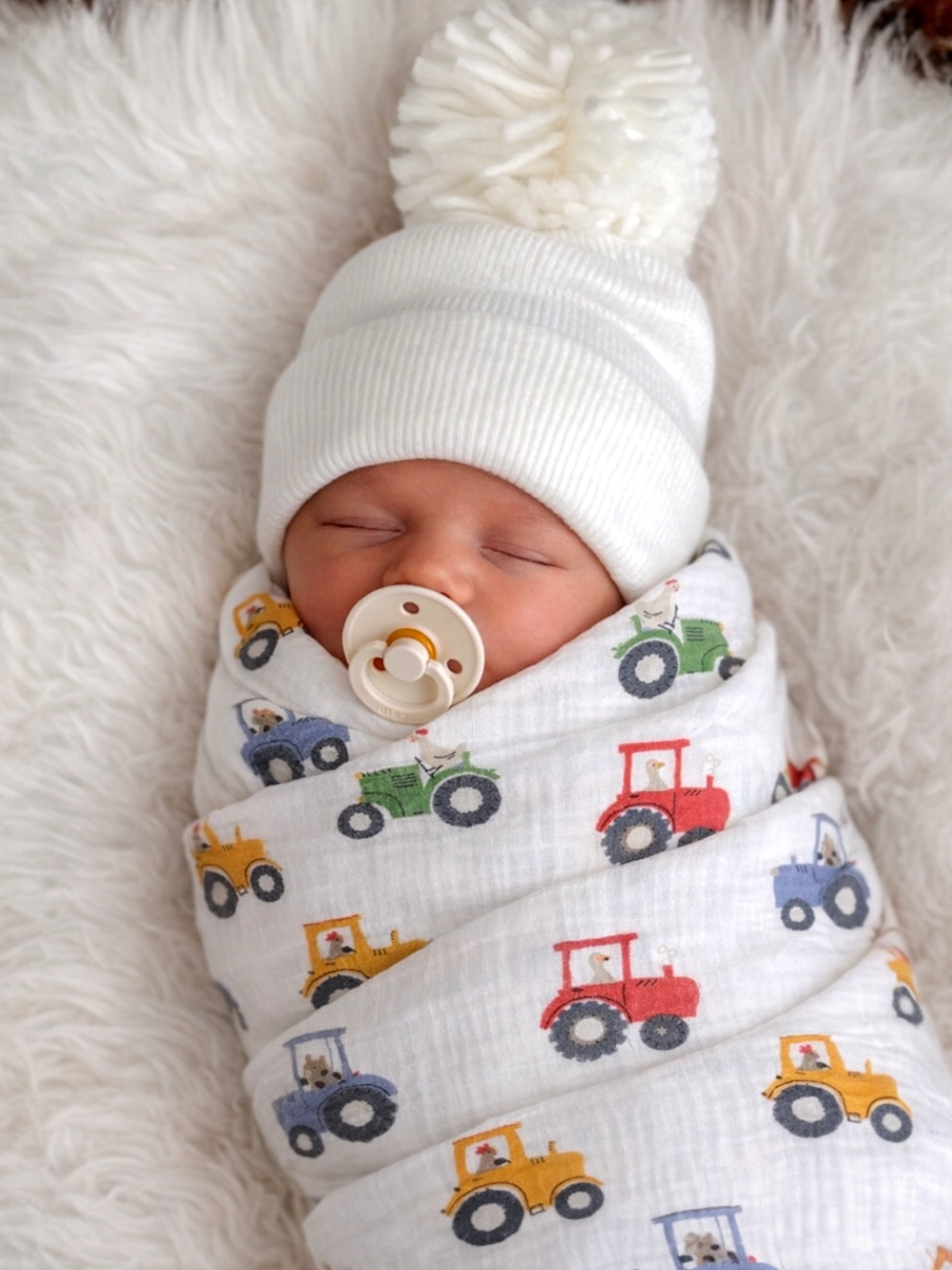Newborn baby sleeping in a white hat with pom pom, swaddled in a tractor-patterned blanket on a soft surface.