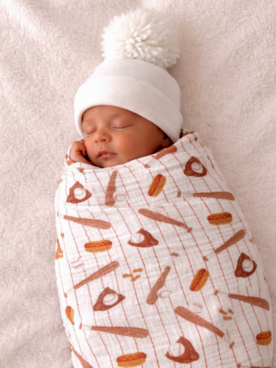 Sleeping baby swaddled in a patterned blanket with food items, wearing a white beanie with a pom-pom, on a soft surface.
