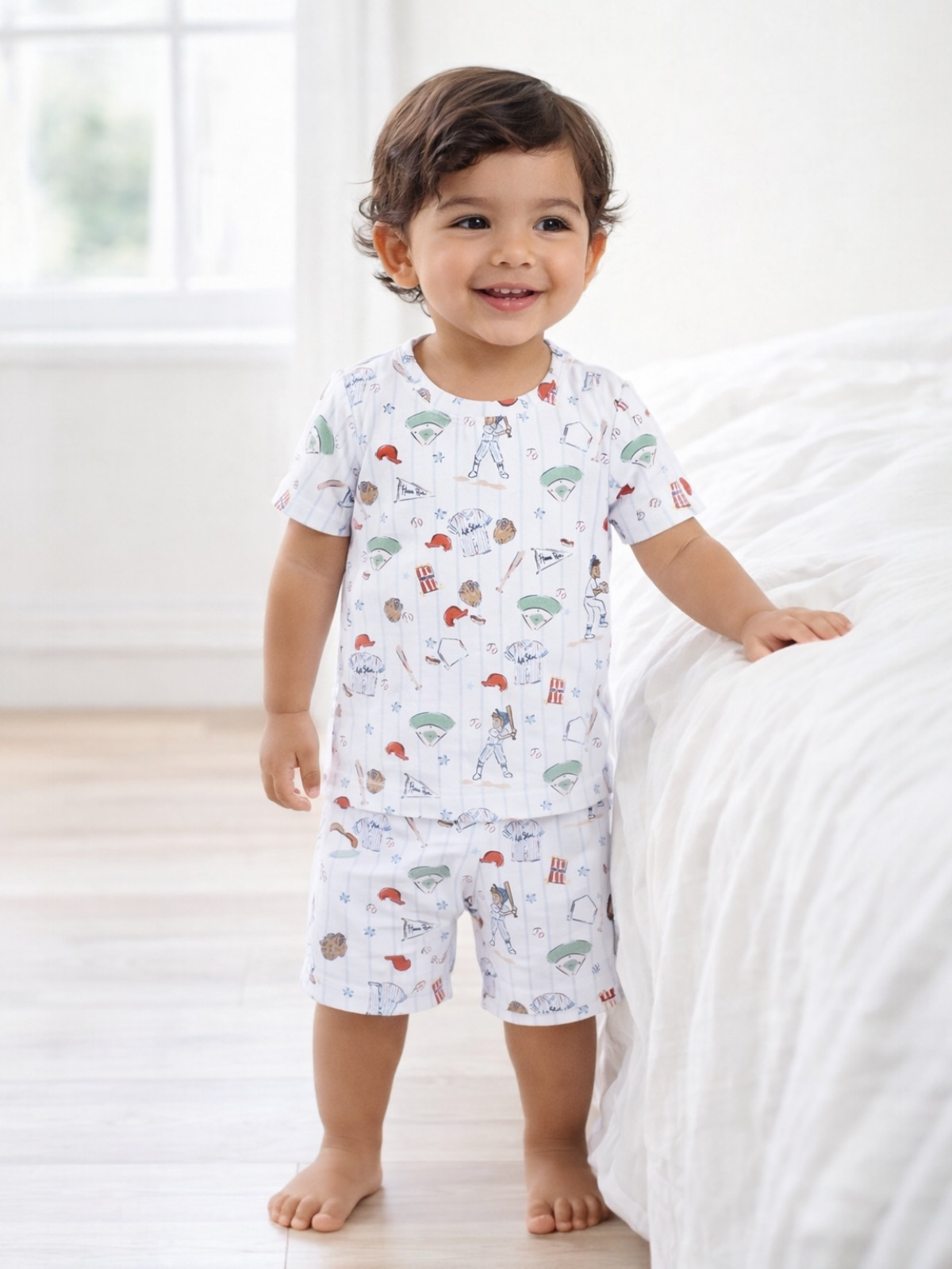 Smiling toddler in playful pajamas, standing by a bed in a bright, airy room.