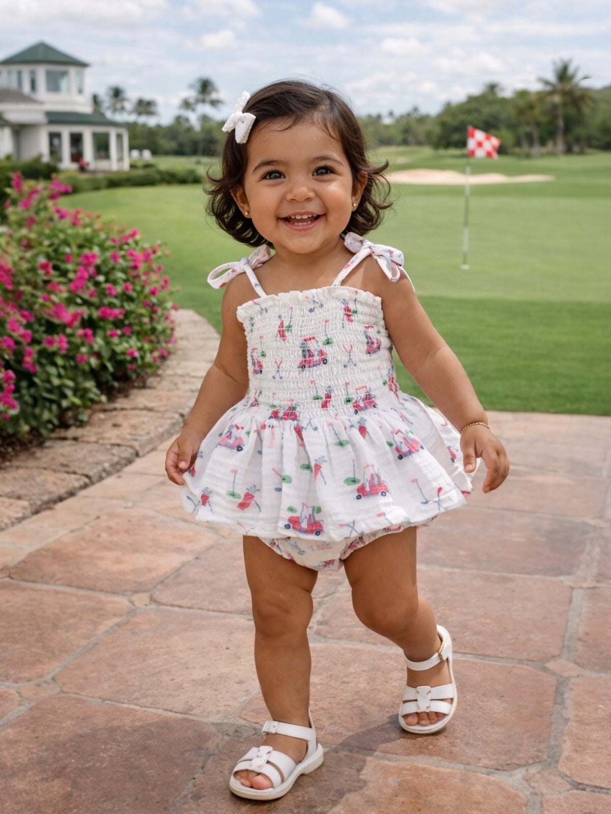 Smiling toddler in a floral outfit walking on a paved path, with a golf course and flowers in the background.