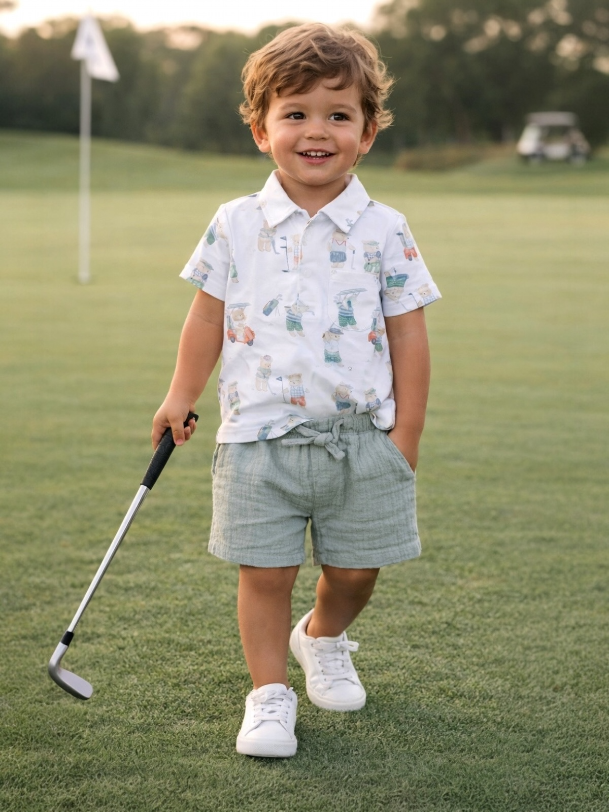 Smiling boy in a patterned shirt and shorts, holding a golf club on a green golf course.