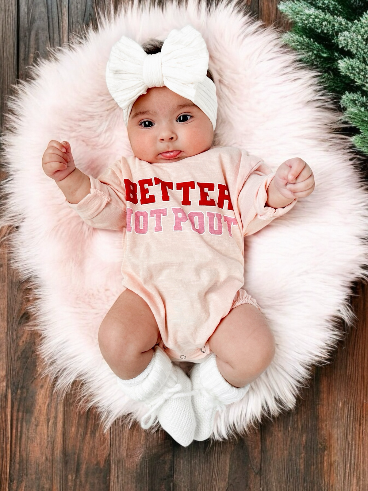 A smiling baby in a pink onesie with "BETTER NOT POUT" text, lying on a fluffy rug with a large bow headband.