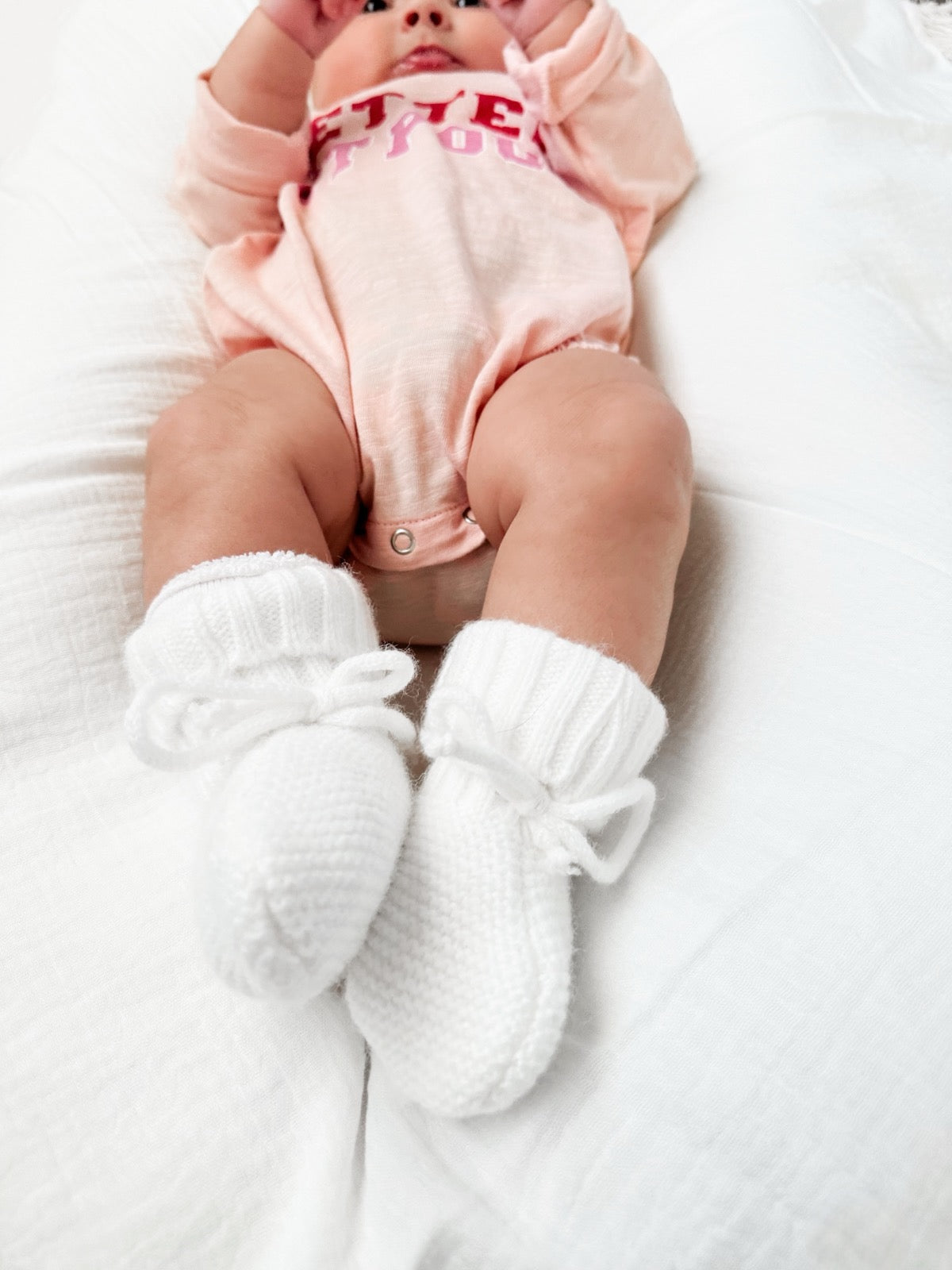 Baby wearing a pink onesie and white knitted booties, lying on a soft blanket.