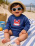 Smiling child in sunglasses, wearing a crab-themed outfit, sitting on a beach towel by the ocean.