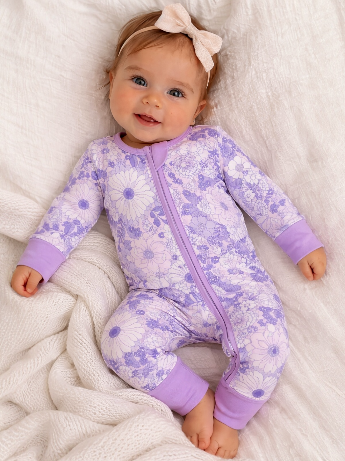 Smiling baby in a floral lavender onesie with a matching headband, lying on a cozy white blanket.