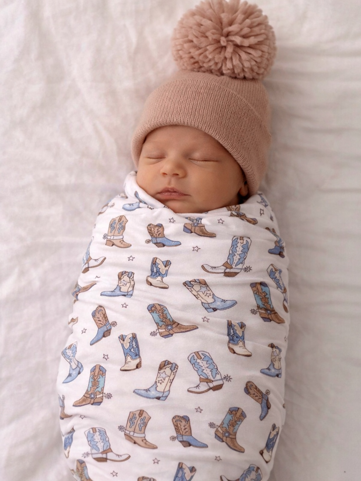 Sleeping baby wrapped in boot-patterned blanket, wearing a beige hat with a pompom on a soft white surface.