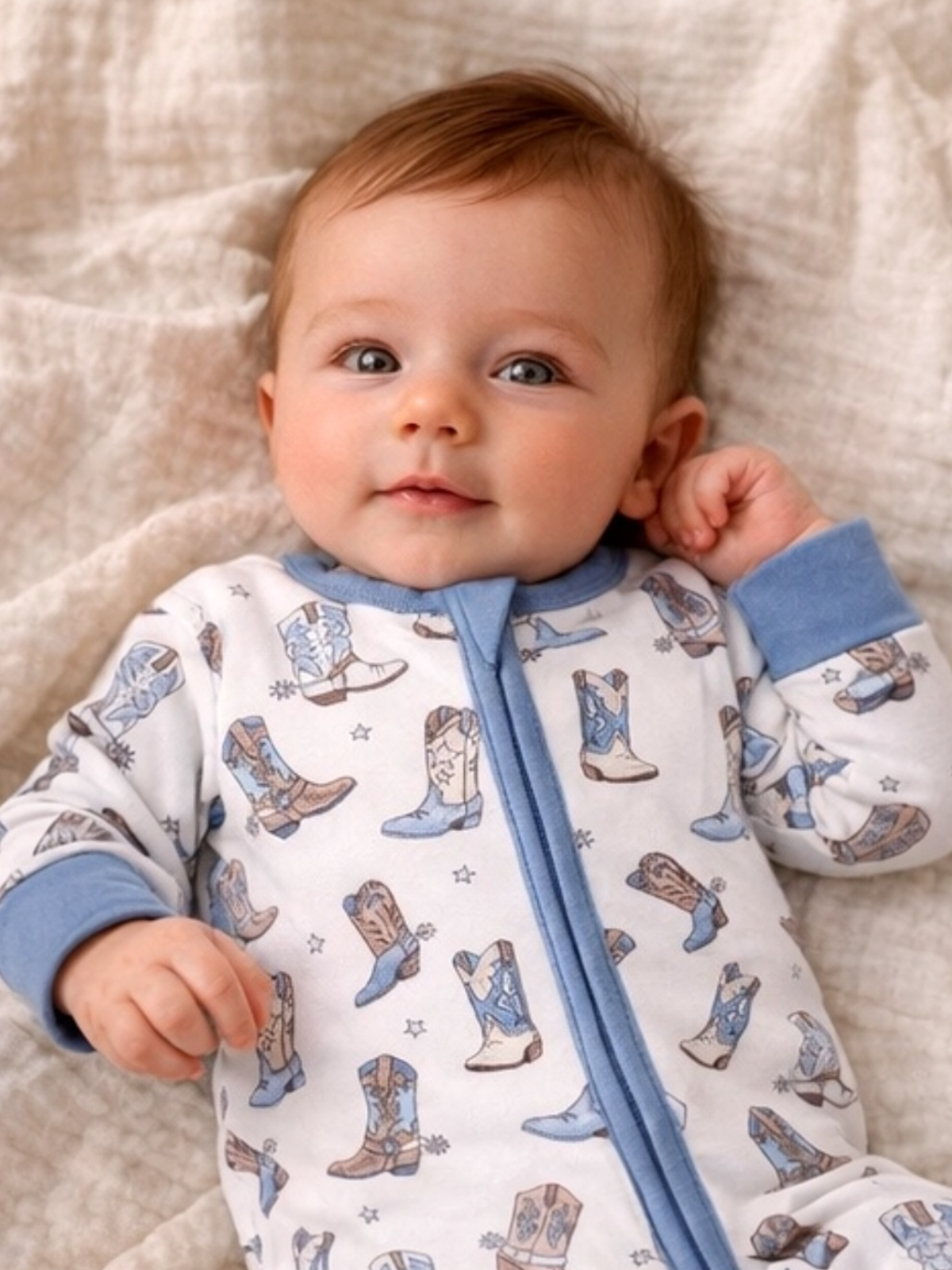 Infant in a cowboy boot-printed onesie lies on a soft blanket, smiling and looking at the camera.