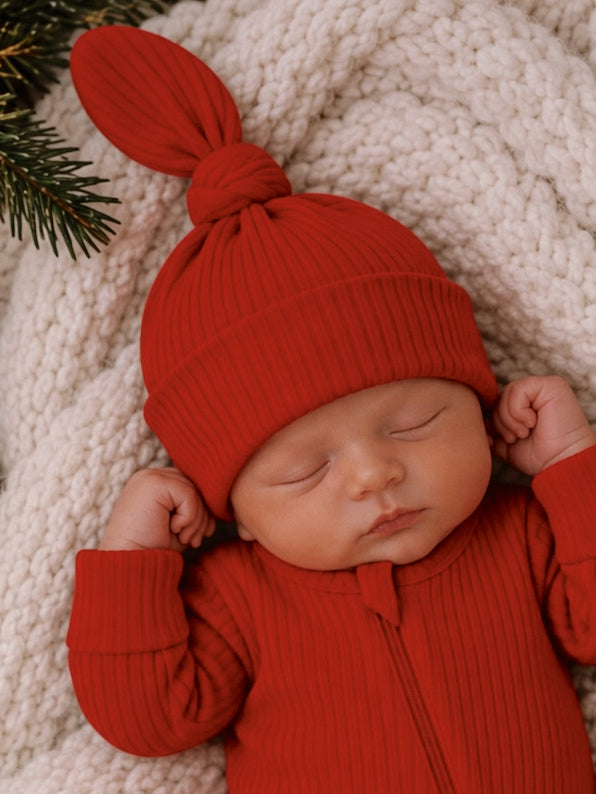 A peaceful sleeping baby in a red ribbed outfit and hat on a cozy cream blanket.