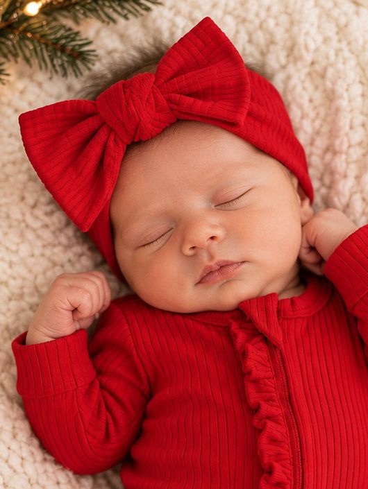 Sleeping baby in a red outfit with a large bow, resting on a cozy blanket with festive decorations.