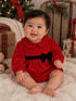 Baby in a red velvet dress with black bow, sitting on a fluffy rug, smiling amidst holiday decorations.