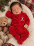 Sleeping baby in a red onesie, cuddled with a teddy bear, surrounded by festive decorations and cozy blankets.