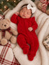Infant in red velvet outfit sleeping peacefully beside a teddy bear, surrounded by festive decorations and blankets.