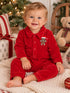 Smiling toddler in red pajamas with a gingerbread man on the chest, seated on a cozy blanket near Christmas decor.