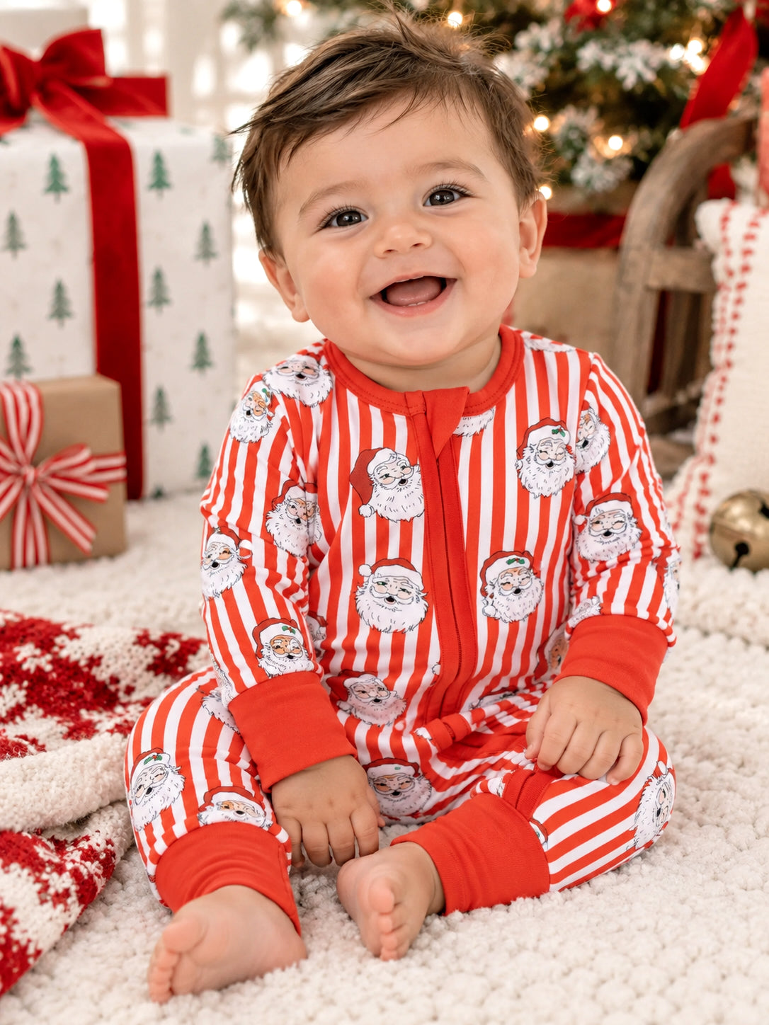 Toddler in festive striped pajamas with Santa faces, smiling on a soft rug surrounded by holiday gifts.