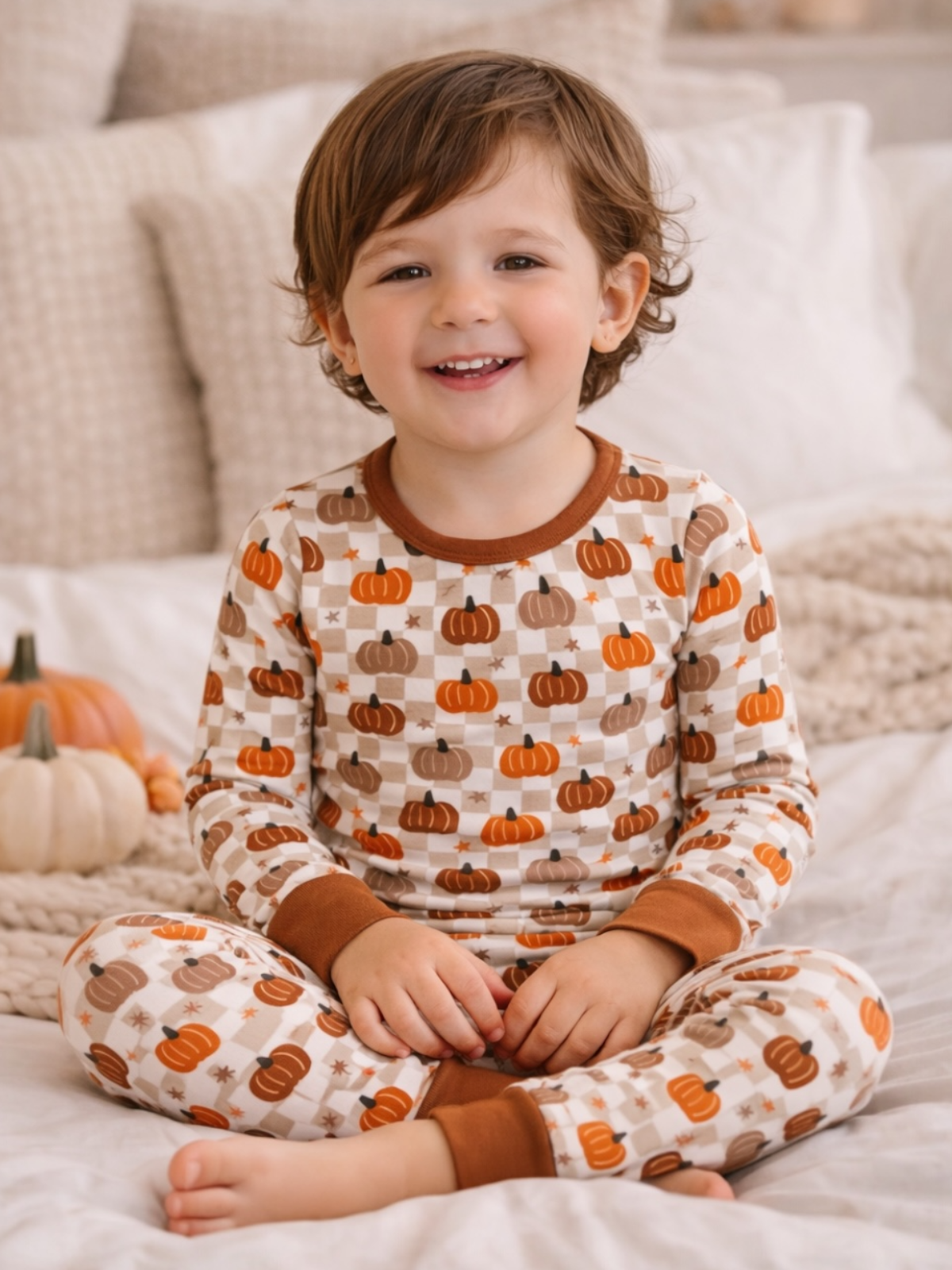 Smiling child in pumpkin-patterned pajamas, sitting on a cozy bed with soft blankets and fall decor.