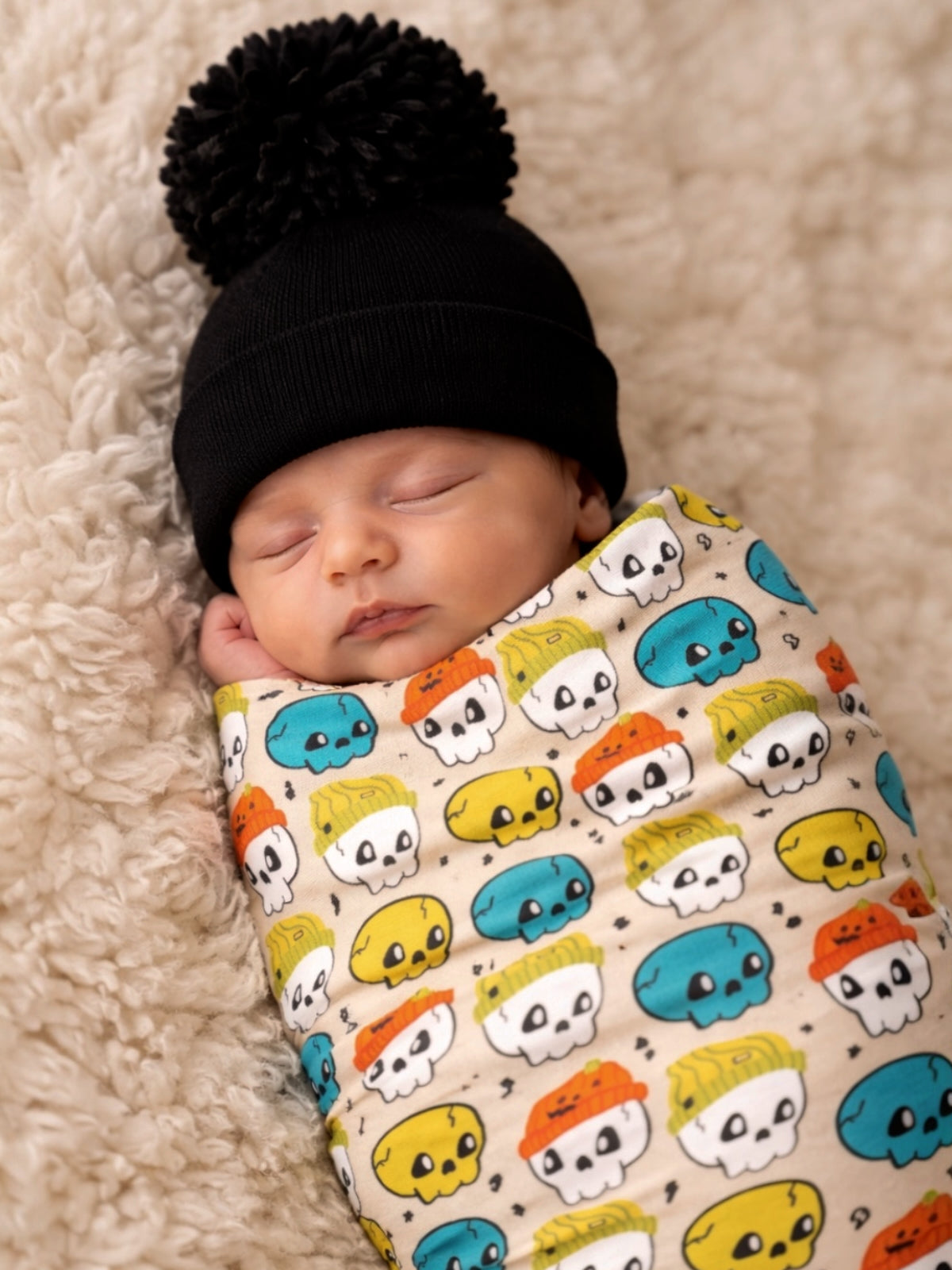 Newborn baby sleeping on a fluffy blanket, swaddled in a skull-patterned fabric and wearing a black pom-pom hat.