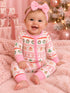Smiling baby in a festive onesie with holiday patterns, sitting on pink fur blanket with Christmas tree and gifts.