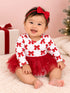 Smiling baby girl in a red bow dress and headband, sitting on a fluffy blanket with Christmas decor in the background.