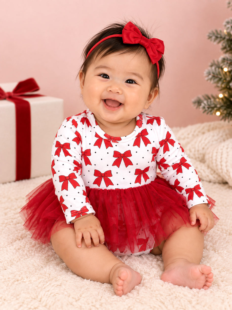 Smiling baby girl in a red bow dress and headband, sitting on a fluffy blanket with Christmas decor in the background.