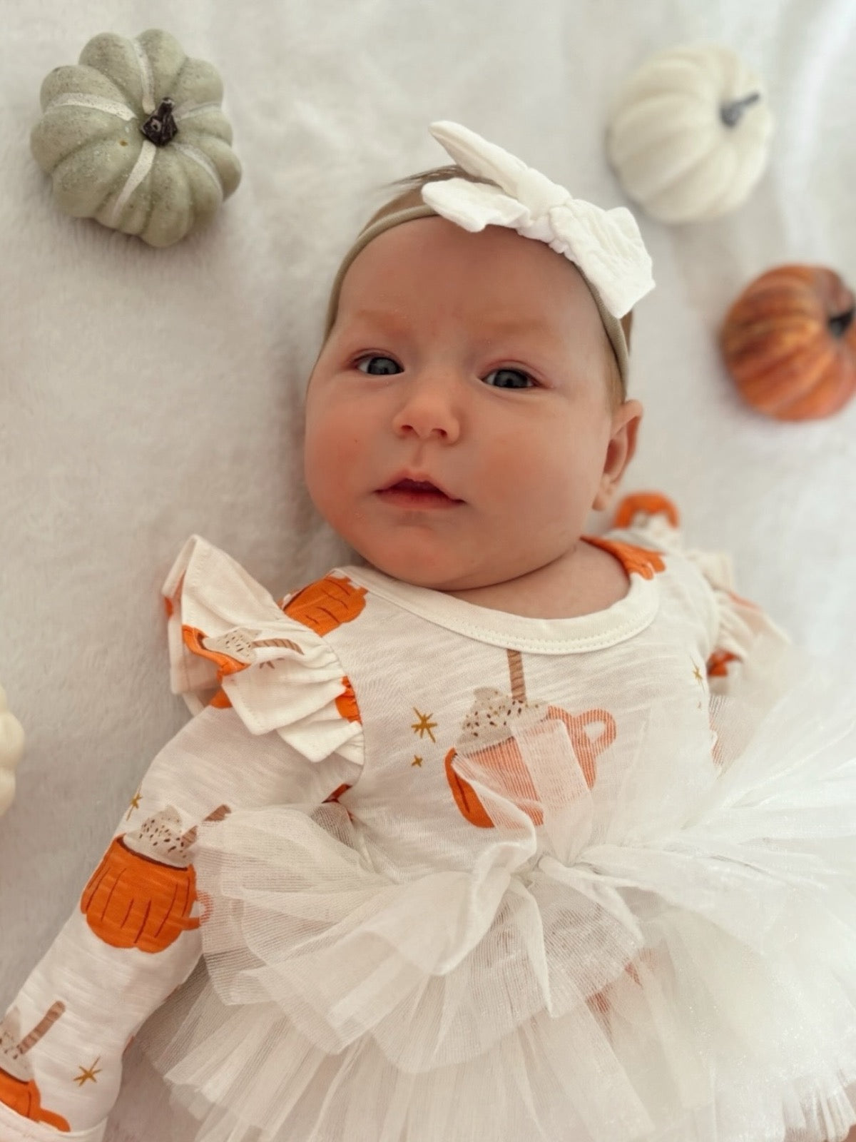 Baby girl in a pumpkin-themed outfit with tulle, surrounded by small decorative pumpkins.