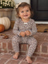 Smiling toddler in leopard print outfit sits on brick steps, surrounded by pumpkins and flowers for fall decor.