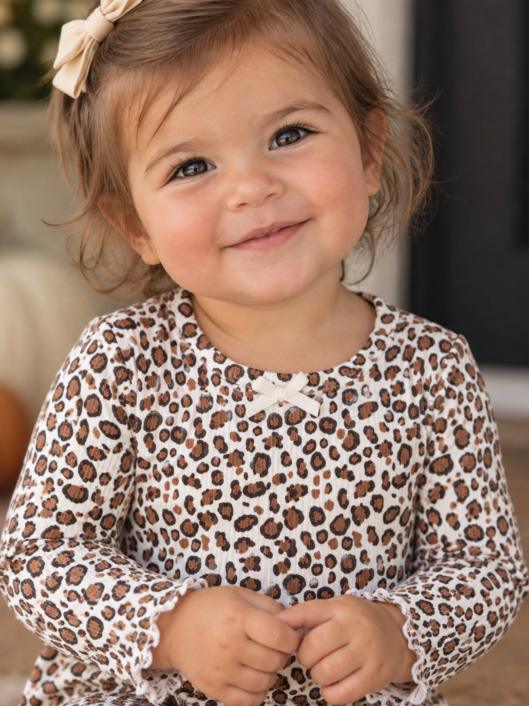 Smiling toddler in a leopard print outfit with a bow, sitting and holding hands, against a neutral background.