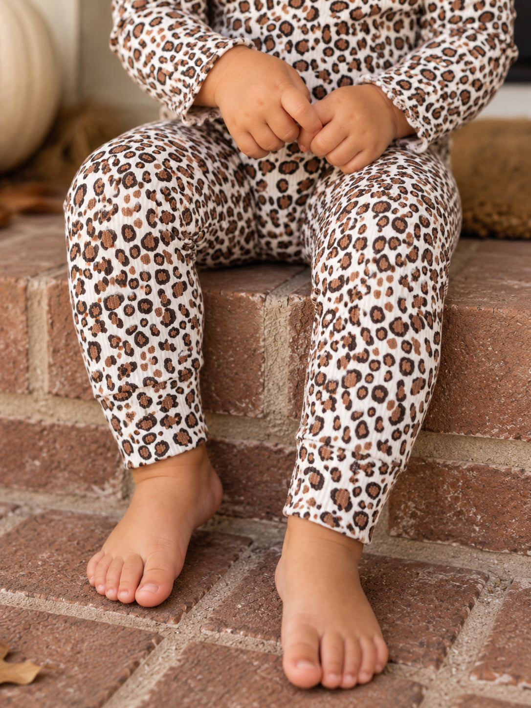 Child in leopard print pajamas sitting on brick steps with bare feet, hands clasped on knees.
