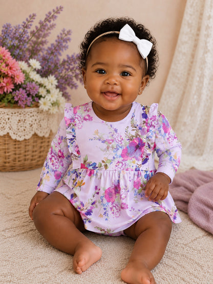 Smiling baby girl in a floral dress and white bow, sitting on a soft surface with flowers in a basket nearby.