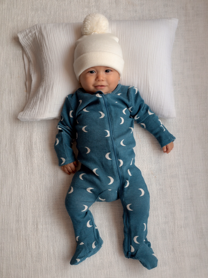 Smiling baby in a blue moon-patterned onesie and white pom-pom hat, lying on a pillow against a textured background.