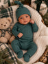 Infant in a green outfit with a matching hat, cuddling a teddy bear among holiday decor and wrapped gifts.