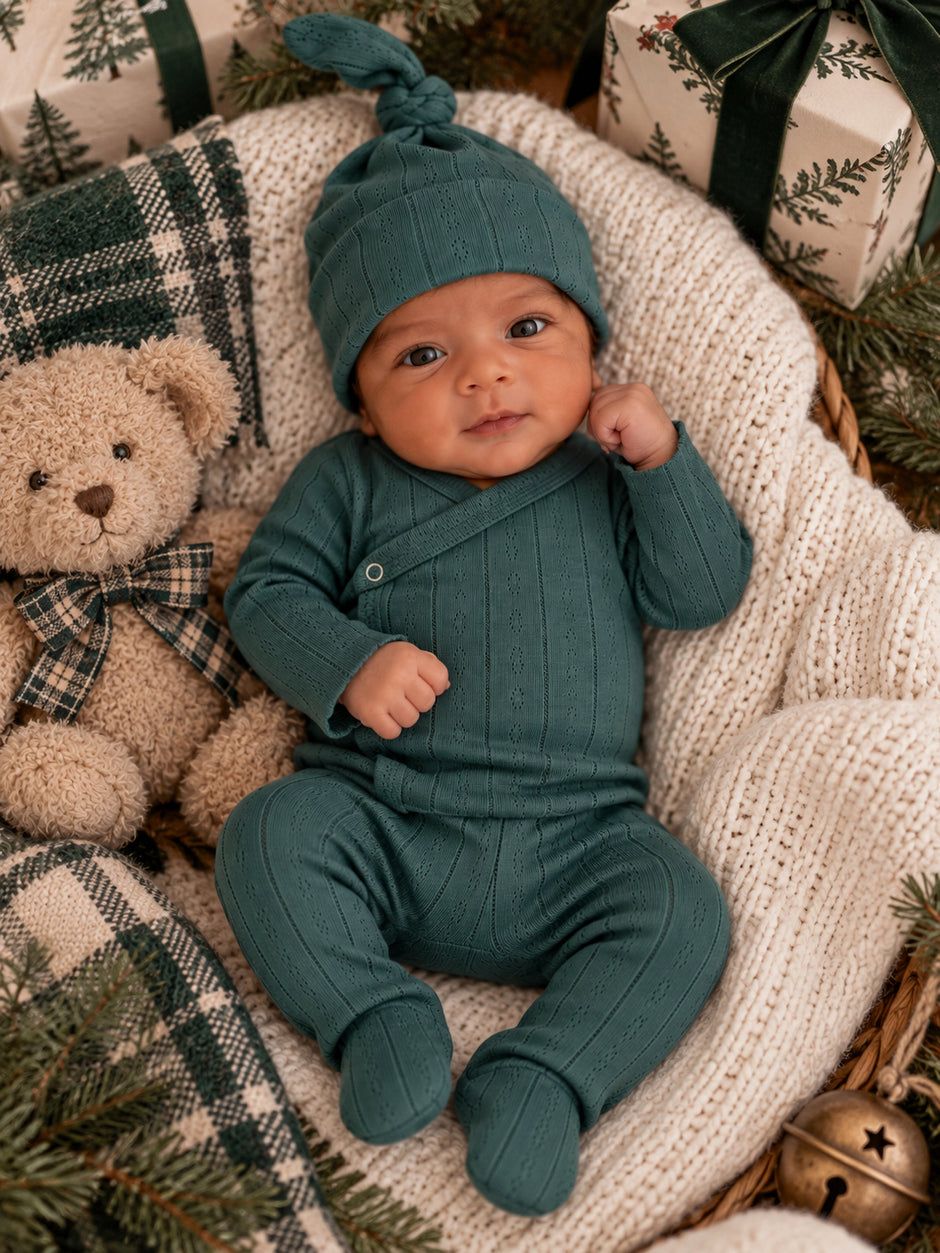 Infant in a green outfit with a matching hat, cuddling a teddy bear among holiday decor and wrapped gifts.