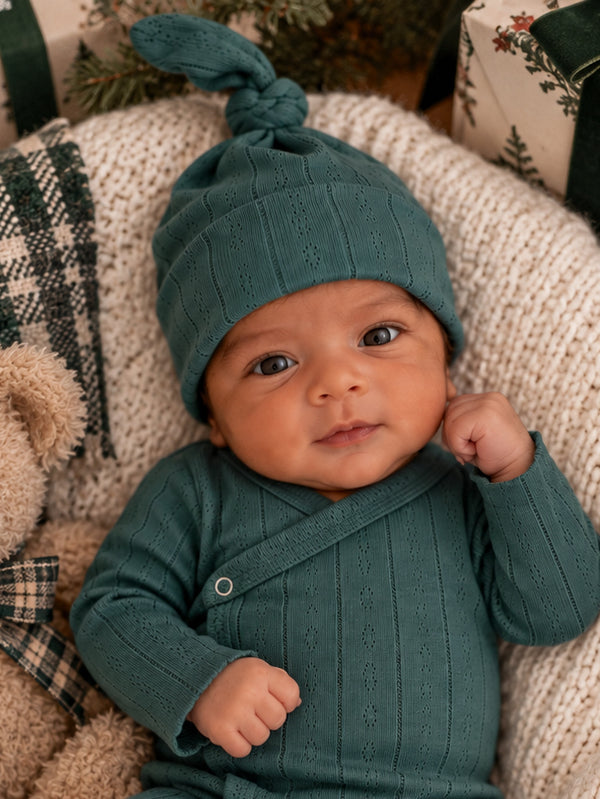 Smiling baby in a teal outfit and matching hat, resting on a cozy blanket with festive decorations in the background.