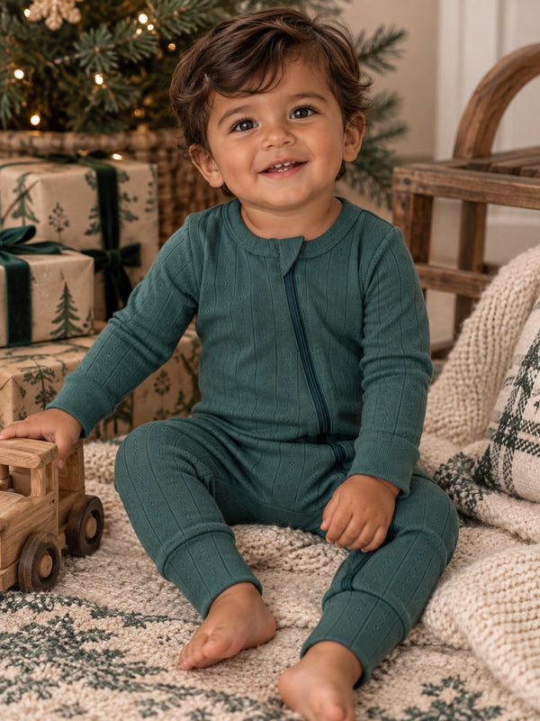 Smiling toddler in green pajamas sitting on a cozy blanket, surrounded by holiday decorations and wooden toys.