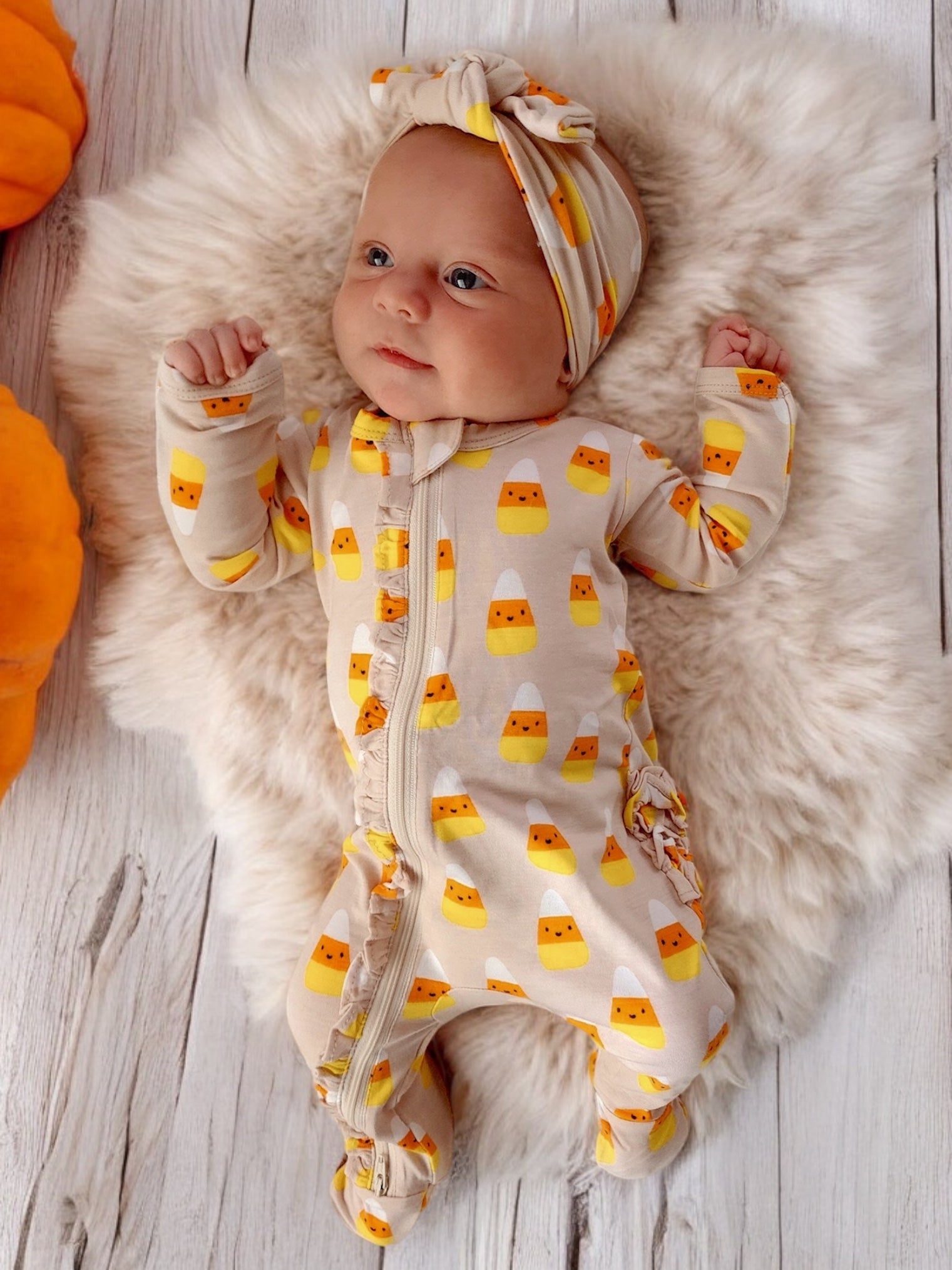 Cute baby in candy corn-patterned outfit lying on a fluffy blanket, surrounded by pumpkins.