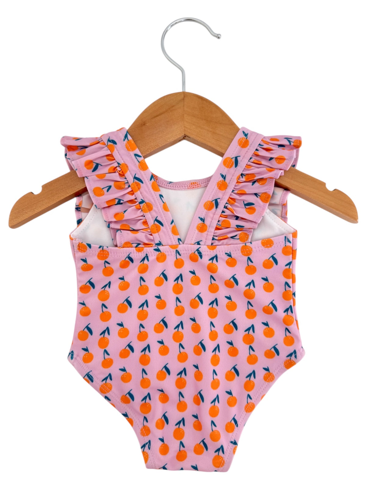 Pink one-piece swimsuit with orange fruit pattern and ruffled straps, hanging on a wooden hanger.