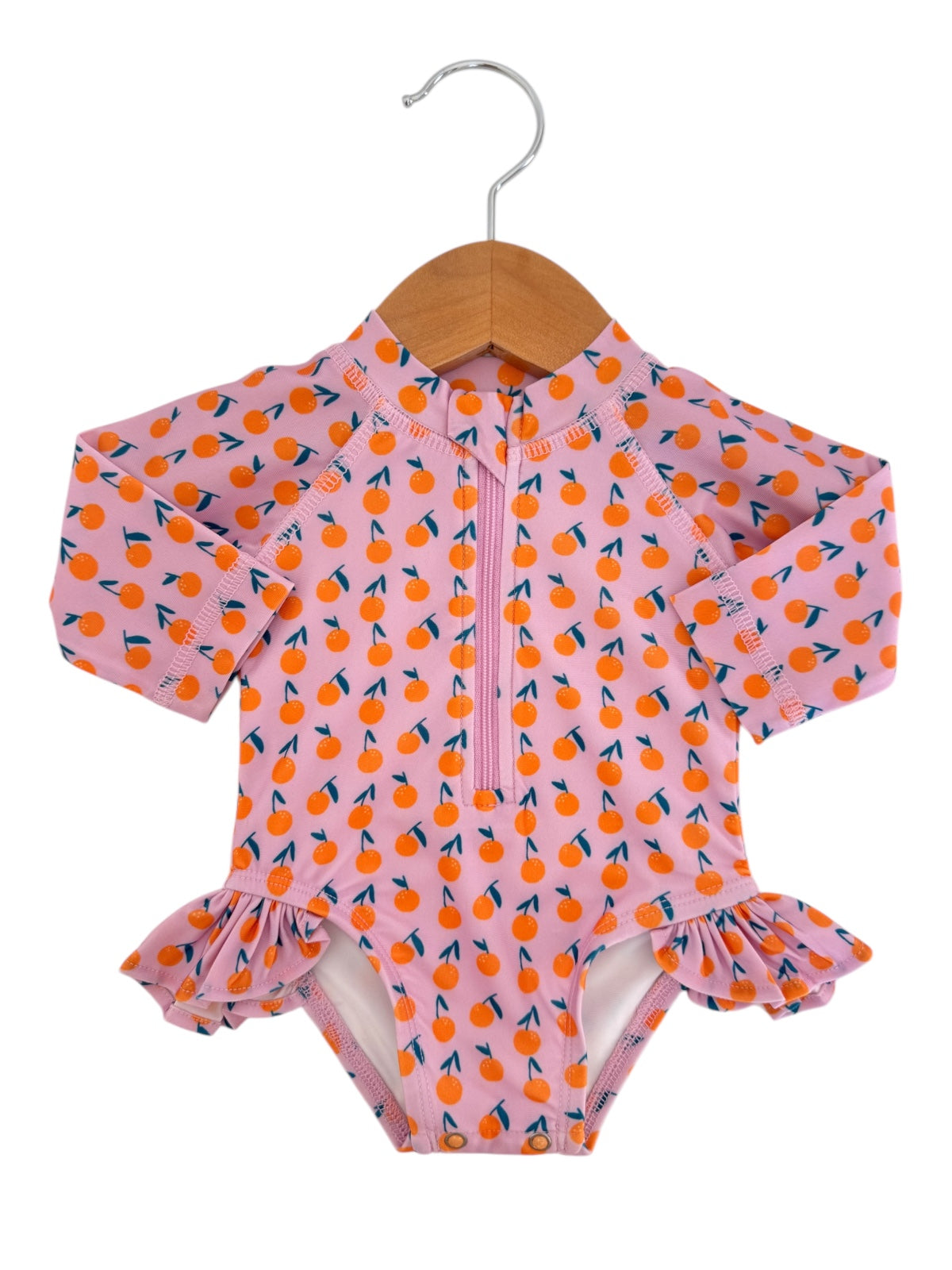 Pink swimsuit with orange fruit pattern, long sleeves, and ruffles, displayed on a wooden hanger.