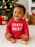 Smiling baby in a red "Santa Baby" outfit sitting on a cozy rug with a Christmas tree in the background.