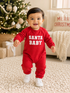 Smiling baby in red "Santa Baby" outfit, walking on carpet beside a decorated Christmas tree.