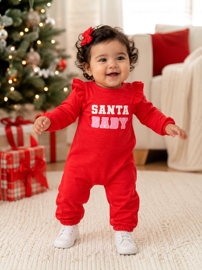 Smiling baby in red "SANTA BABY" outfit stands near a decorated Christmas tree with presents.