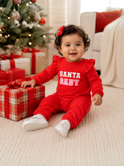 Smiling baby in festive red outfit sitting near Christmas tree and gift boxes, celebrating the holiday season.