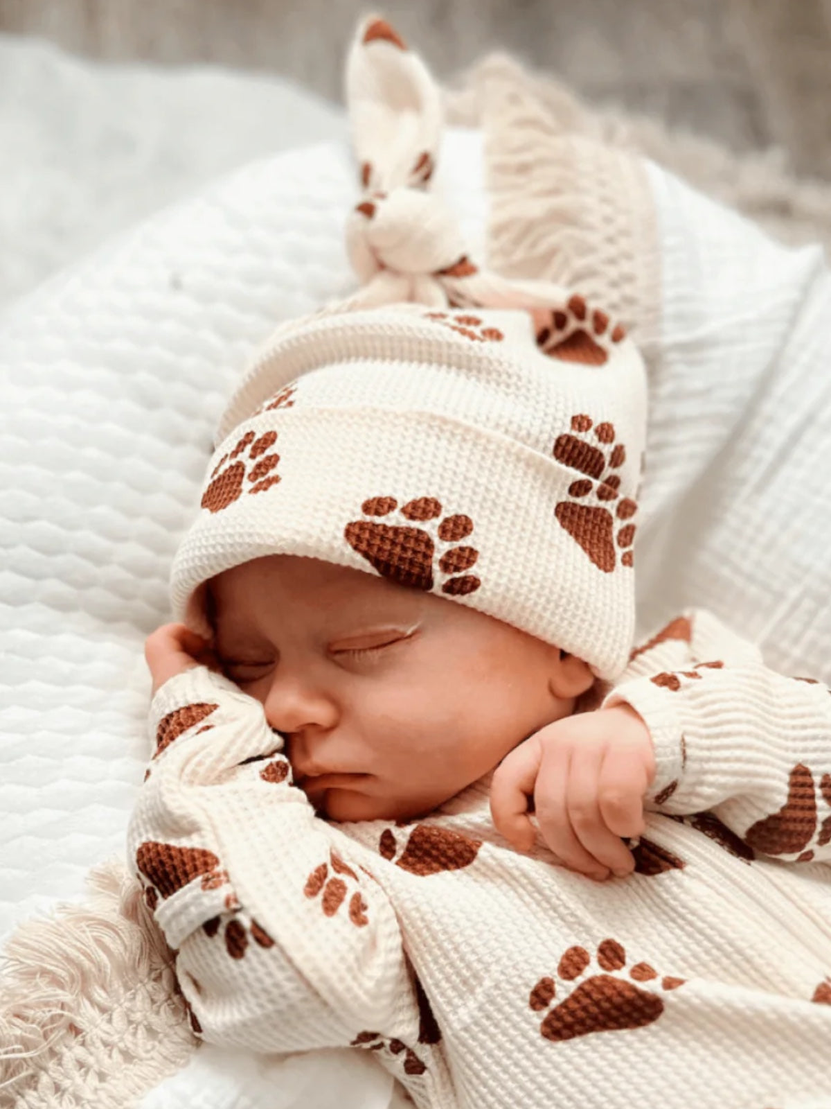 Sleeping baby in cozy outfit with brown paw prints, wearing a matching hat, on a textured blanket.