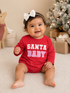 Baby in a red "Santa Baby" onesie sits on a soft rug, smiling, with a white bow in her hair and holiday decorations nearby.