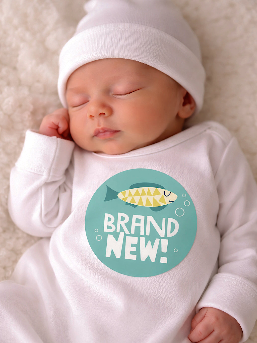 Sleeping baby in a white onesie with "BRAND NEW!" text and fish graphic, resting on a fluffy blanket.