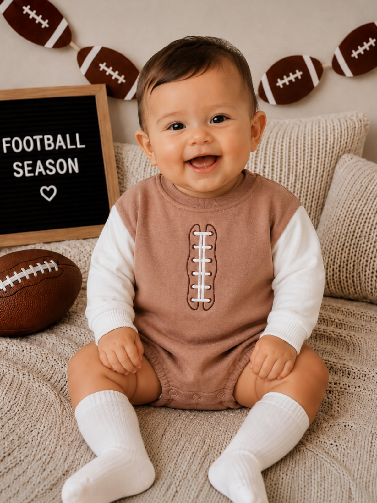 Smiling baby in football-themed outfit, sitting on a cozy blanket near a football and "Football Season" sign.